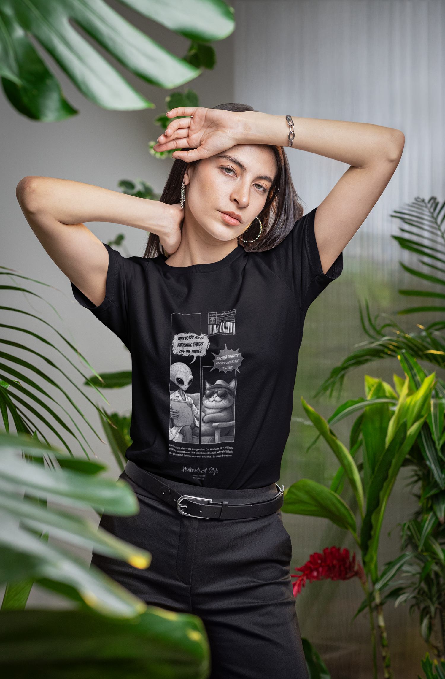 Woman standing in a lush indoor plant setting, wearing a black t-shirt featuring a comic-style design of an alien and a smug cat. The alien asks, “Why do you keep knocking things off the table?” and the cat replies, “Because gravity needs constant testing.” The artwork includes barcode graphics and small scientific-style print. She poses with one arm resting on her head, framed by large green tropical leaves in a soft-lit modern interior.