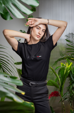 A stylish woman poses confidently among tropical plants, wearing a black t-shirt with a minimalist "Istanbul TR" design. The shirt features the Galata Tower icon, modern typography, and a red-white bar symbolizing the Turkish flag.