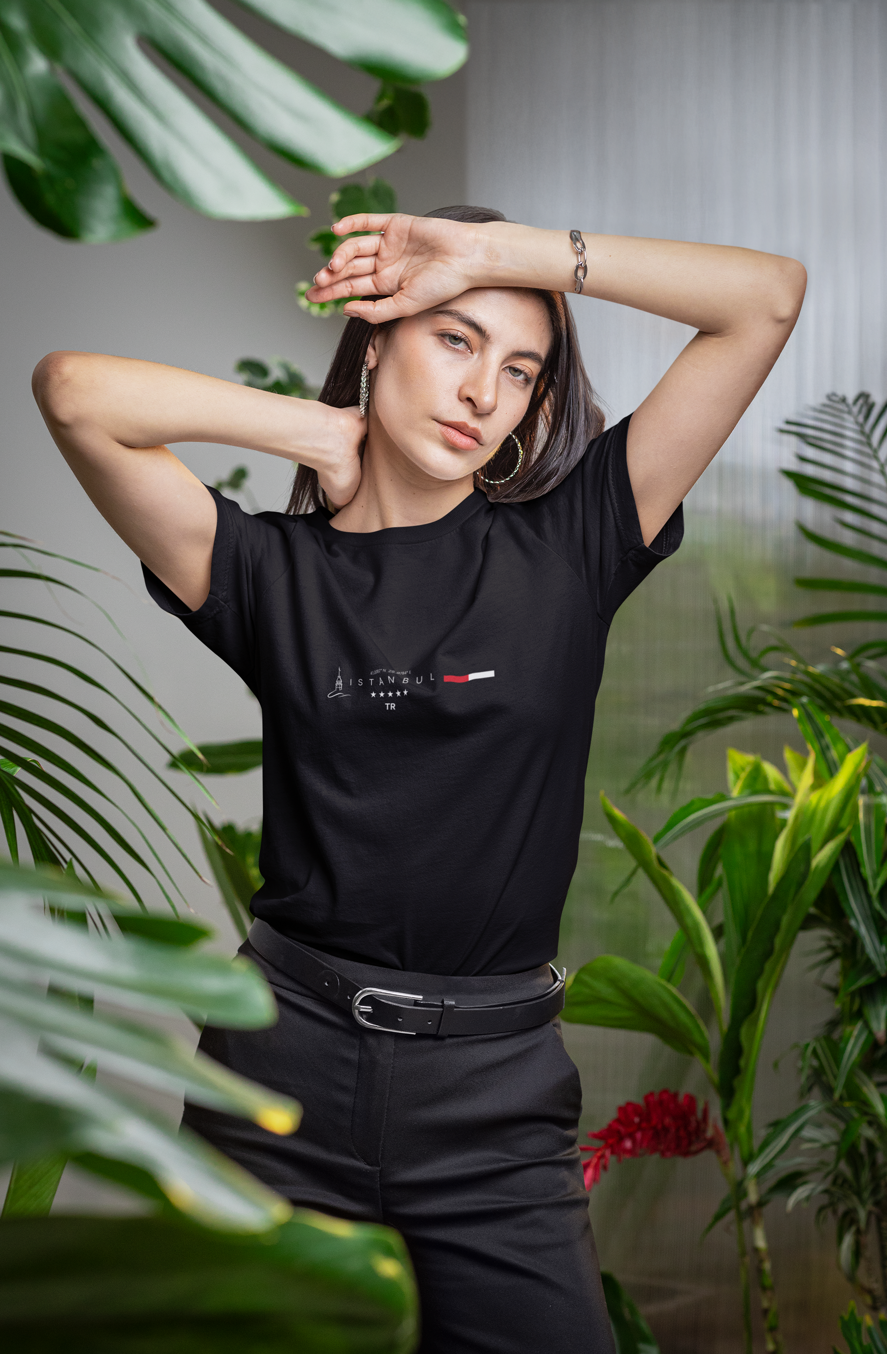 A stylish woman poses confidently among tropical plants, wearing a black t-shirt with a minimalist "Istanbul TR" design. The shirt features the Galata Tower icon, modern typography, and a red-white bar symbolizing the Turkish flag.