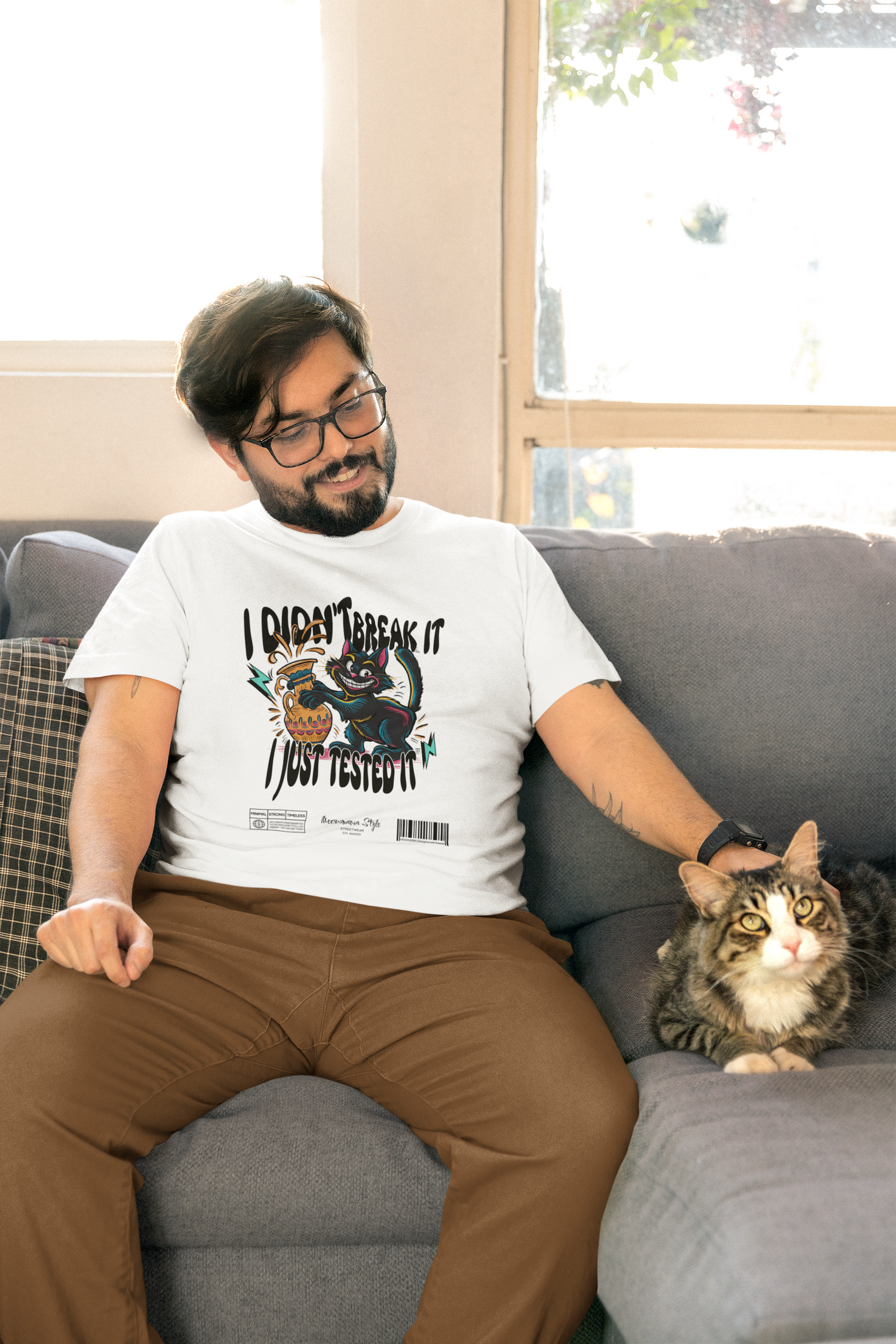 Man sitting casually on a grey couch in a sunlit living room, wearing a white t-shirt with a playful cartoon design of a mischievous cat and the text “I Didn’t Break It, I Just Tested It.” A tabby cat lies beside him, matching the shirt’s vibe.