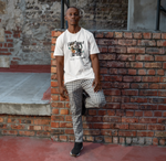 Man standing casually against a brick wall in an urban setting. He wears a white t-shirt with a playful design of a mischievous black cat knocking over a vase, accompanied by the text “I Didn’t Break It, I Just Tested It.” The scene has a relaxed, street-style vibe with plaid pants and sneakers.