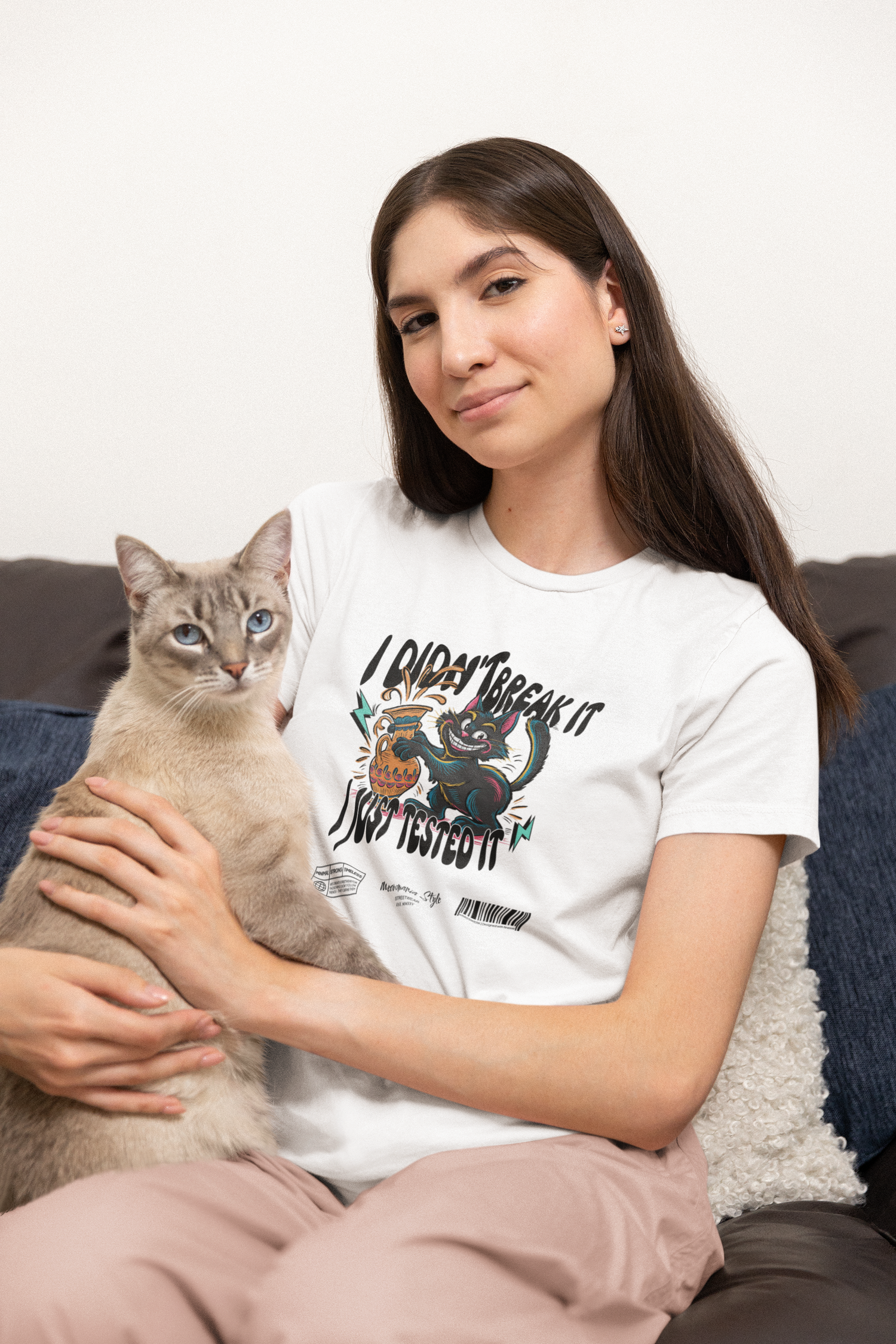 Woman sitting on a dark sofa, gently holding a light gray Siamese cat in her arms. She wears a white t-shirt featuring a cartoon black cat spilling a vase, with the caption “I Didn’t Break It, I Just Tested It.” The scene is cozy, with calm expressions and warm lighting.