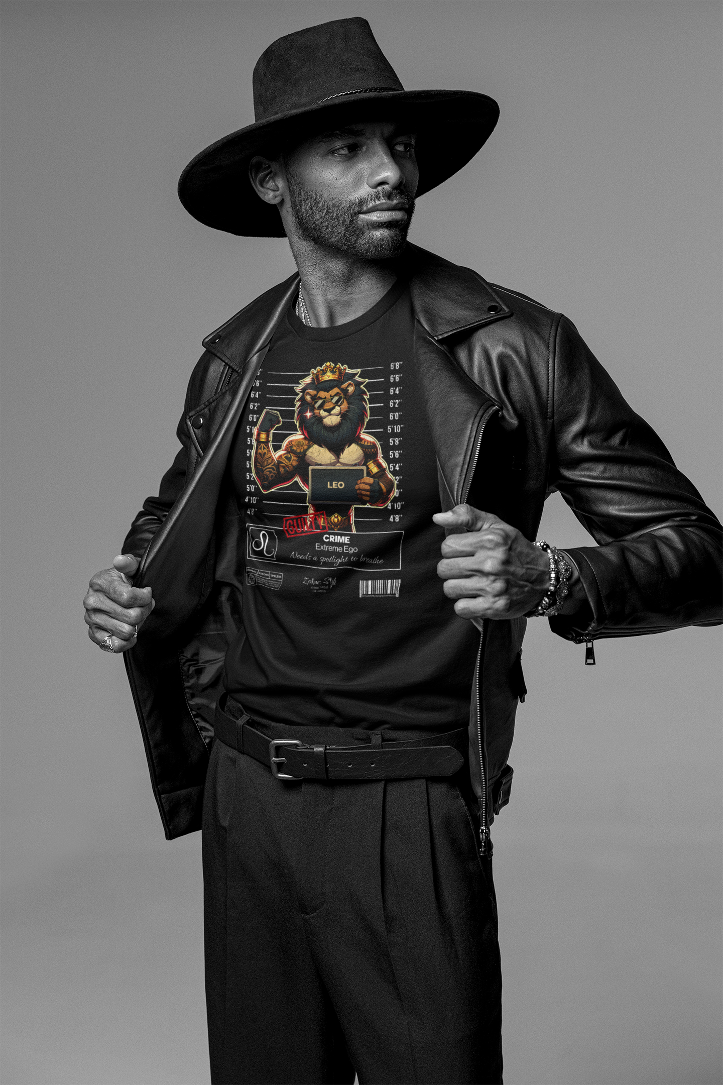 Black and white photo of a confident man revealing his black t-shirt by pulling open his leather jacket. He wears a wide-brimmed hat, multiple bracelets, and dark pleated pants. The t-shirt displays the “LEO” zodiac design — a tattooed lion in a mugshot pose, wearing a crown, flexing a muscular arm, and holding a nameplate. “CRIME: Extreme Ego” is written underneath, stamped with “GUILTY.” Grey studio backdrop. The scene emphasizes bold attitude and dominance.