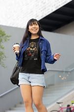 A young woman smiling as she walks down outdoor stairs, wearing a black Leo-themed T-shirt featuring a muscular lion with a crown and tattoos in a mugshot lineup. She holds a coffee cup in one hand and has a black shoulder bag on the other. The T-shirt reads “CRIME: Extreme Ego – Always a spotlight to invade.” She wears light blue denim shorts and an open cobalt corduroy shirt over the tee. The background shows a modern concrete building with stairs, a pink bicycle, and soft natural light.