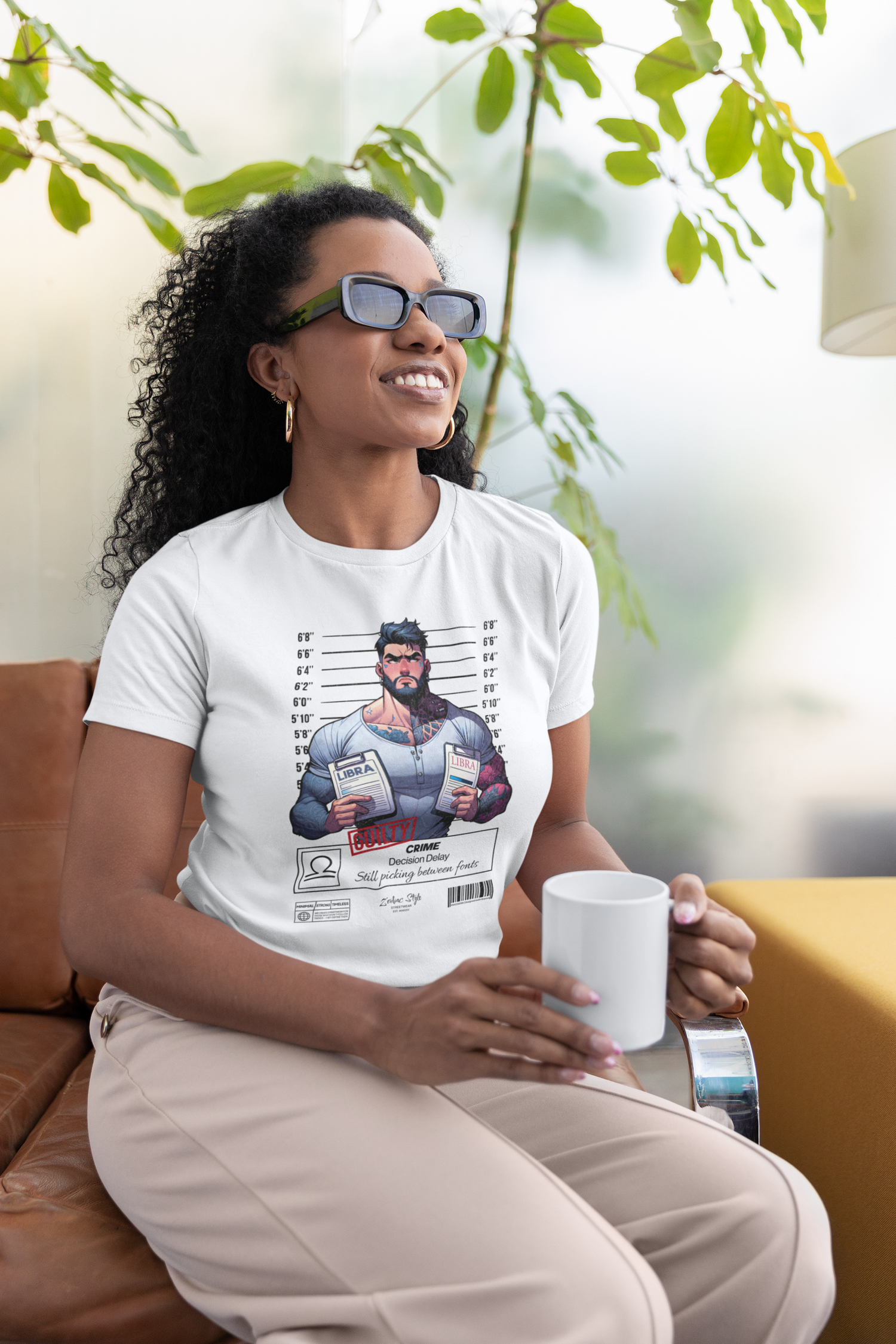 Woman sitting on a couch, holding a mug and wearing the LIBRA t-shirt in white. Bright indoor setting with plants in the background. Shirt design features a muscular man holding two LIBRA folders, labeled with the crime: Decision Delay.