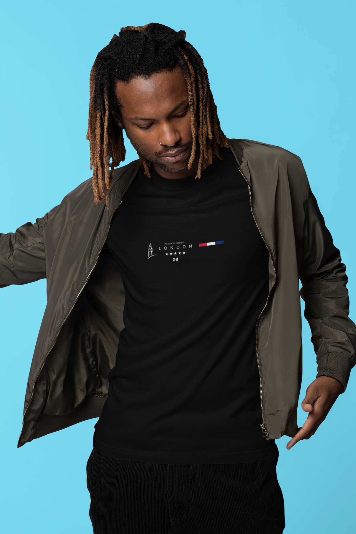 A Black male model with dreadlocks wears the LONDON black t-shirt while opening a green bomber jacket against a vibrant blue background. The design features minimalist city text, coordinates, and a flag motif with five stars.
