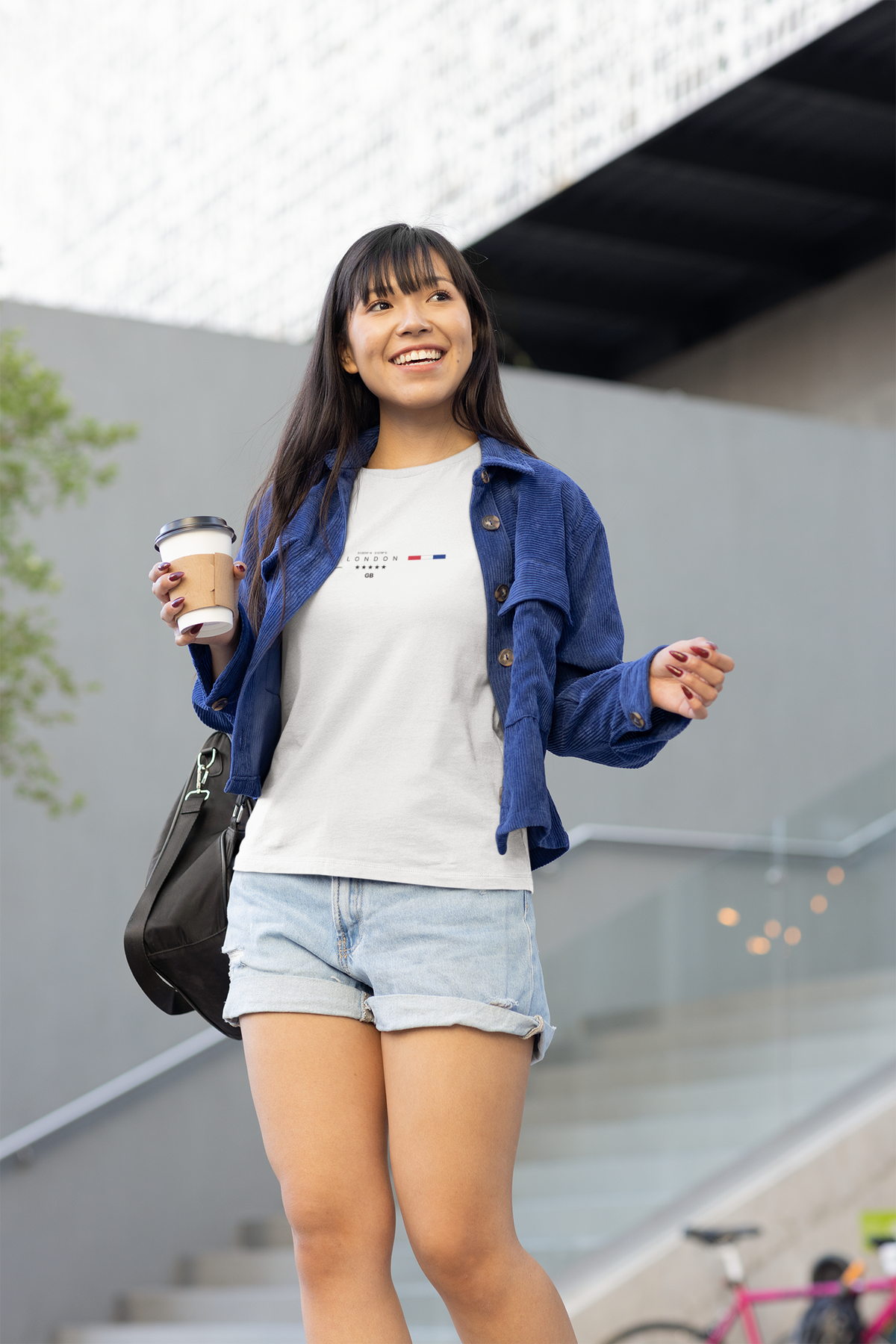 A smiling woman walks outside holding a coffee, wearing the LONDON white t-shirt. The shirt features a minimalist chest design with the Big Ben icon, “LONDON” text, coordinates, five stars, and a red-white-blue stripe. She pairs it with denim shorts and a blue overshirt. Modern architecture and glass stairs form the background.