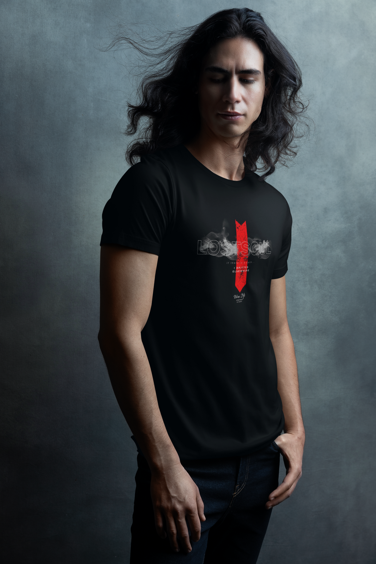 Male model with long flowing hair wearing the “LOSTSOUL” black t-shirt. The design features a smoky translucent title and bold red arrow pointing downward. Shot in a moody, cinematic studio with cool grey textured background.
