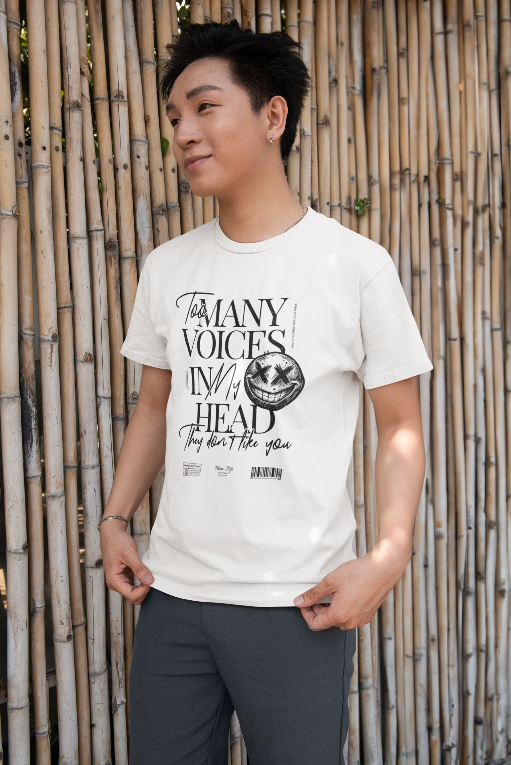 Male model outdoors in front of a bamboo wall, wearing the white “Too Many Voices In My Head” tee. The bold black text and creepy smiley detail pop against the clean background. A subtle rebel vibe.