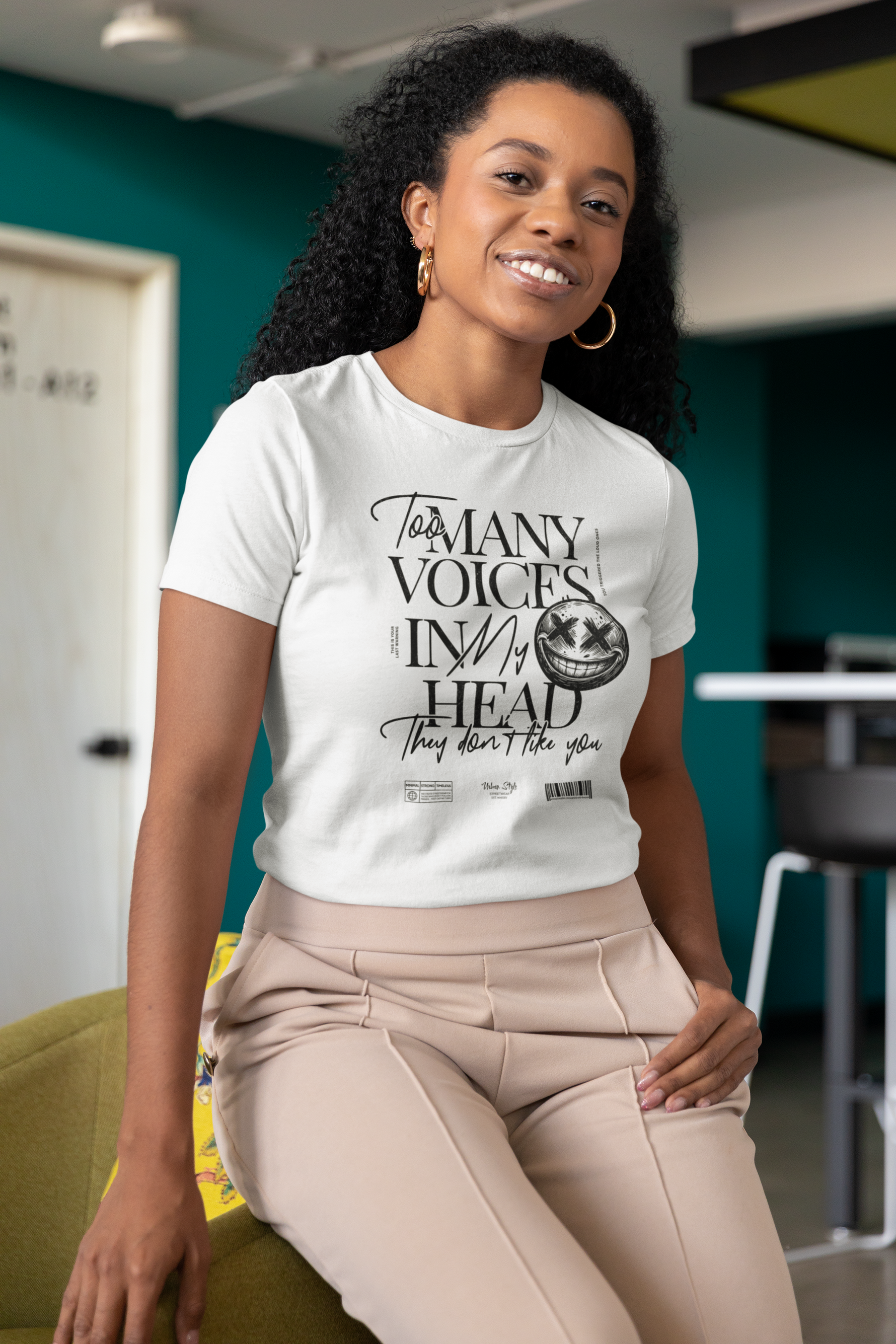 Female model seated in a modern indoor space, smiling confidently in the white “Too Many Voices In My Head” tee. Edgy black typography with a scratched face detail stands out on the soft fabric. Stylish and expressive look.