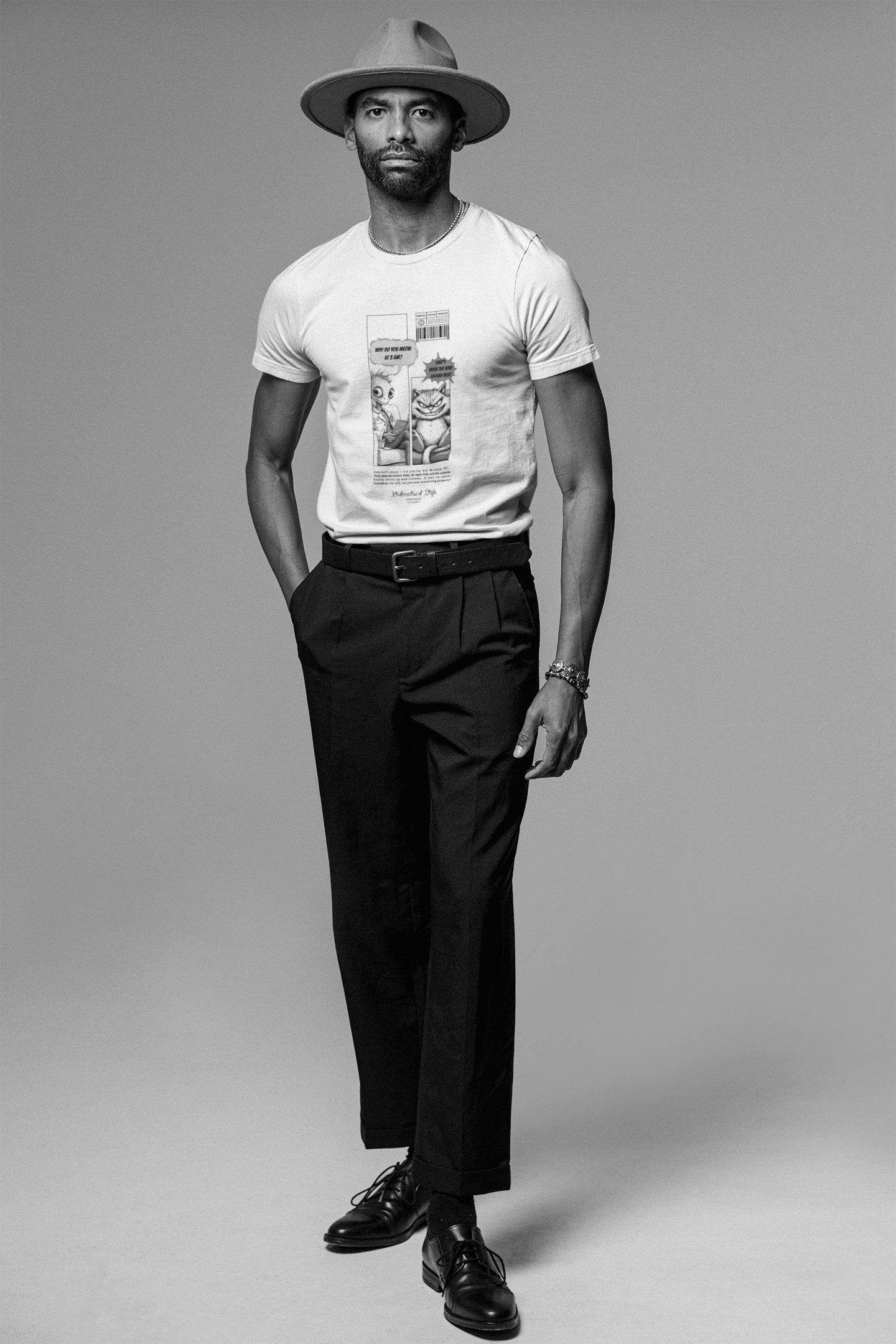 A man in a white t-shirt and wide-brimmed hat stands confidently in a studio. The shirt features a black-and-white comic strip: on the left, an alien asks, “Why do you meow at 3 A.M.?” while on the right, a serious-looking cat responds, “That’s when the void listens best.” The design is centered and vertically framed, styled like an old-school sci-fi cartoon.
