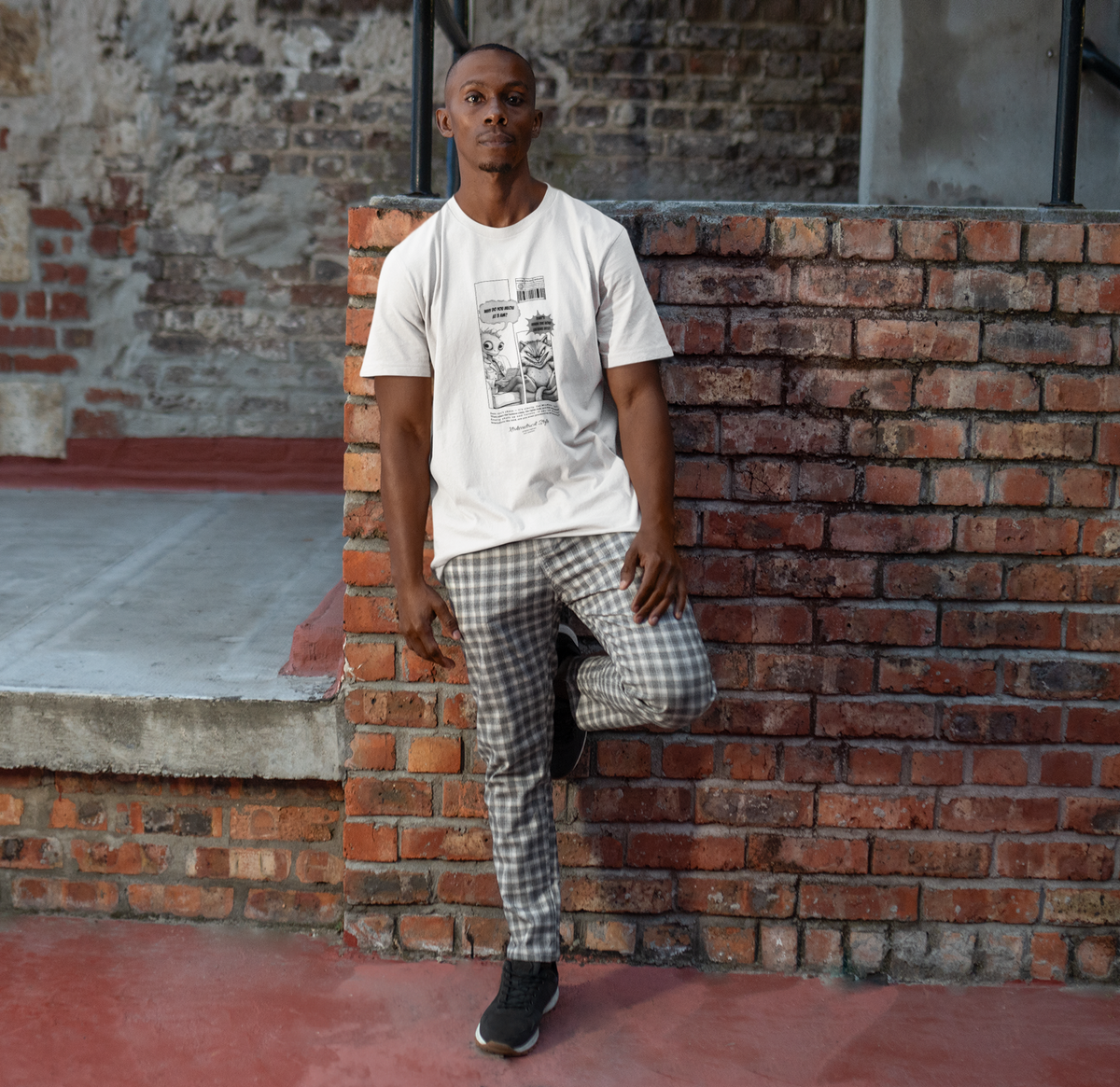 A man leans casually against a brick wall in an urban alleyway, wearing a white t-shirt with a comic-style graphic. The design features a surreal alien asking, “Why do you meow at 3 A.M.?” and a smug-looking cat answering, “That’s when the void listens best.” The scene blends street fashion with dry humor, and the model pairs the tee with checkered pants and sneakers for a laid-back city look.