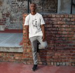 A man leans casually against a brick wall in an urban alleyway, wearing a white t-shirt with a comic-style graphic. The design features a surreal alien asking, “Why do you meow at 3 A.M.?” and a smug-looking cat answering, “That’s when the void listens best.” The scene blends street fashion with dry humor, and the model pairs the tee with checkered pants and sneakers for a laid-back city look.