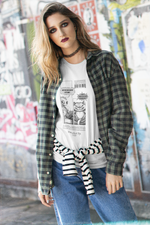 A young woman with bold makeup and loose wavy hair stands against a colorful graffiti wall, wearing a white t-shirt under a green plaid shirt. The t-shirt features a black-and-white comic strip with an alien asking, “Why do you meow at 3 A.M.?” and a smug cat replying, “That’s when the void listens best.” The shirt has a streetwear aesthetic, blending humor and urban style.