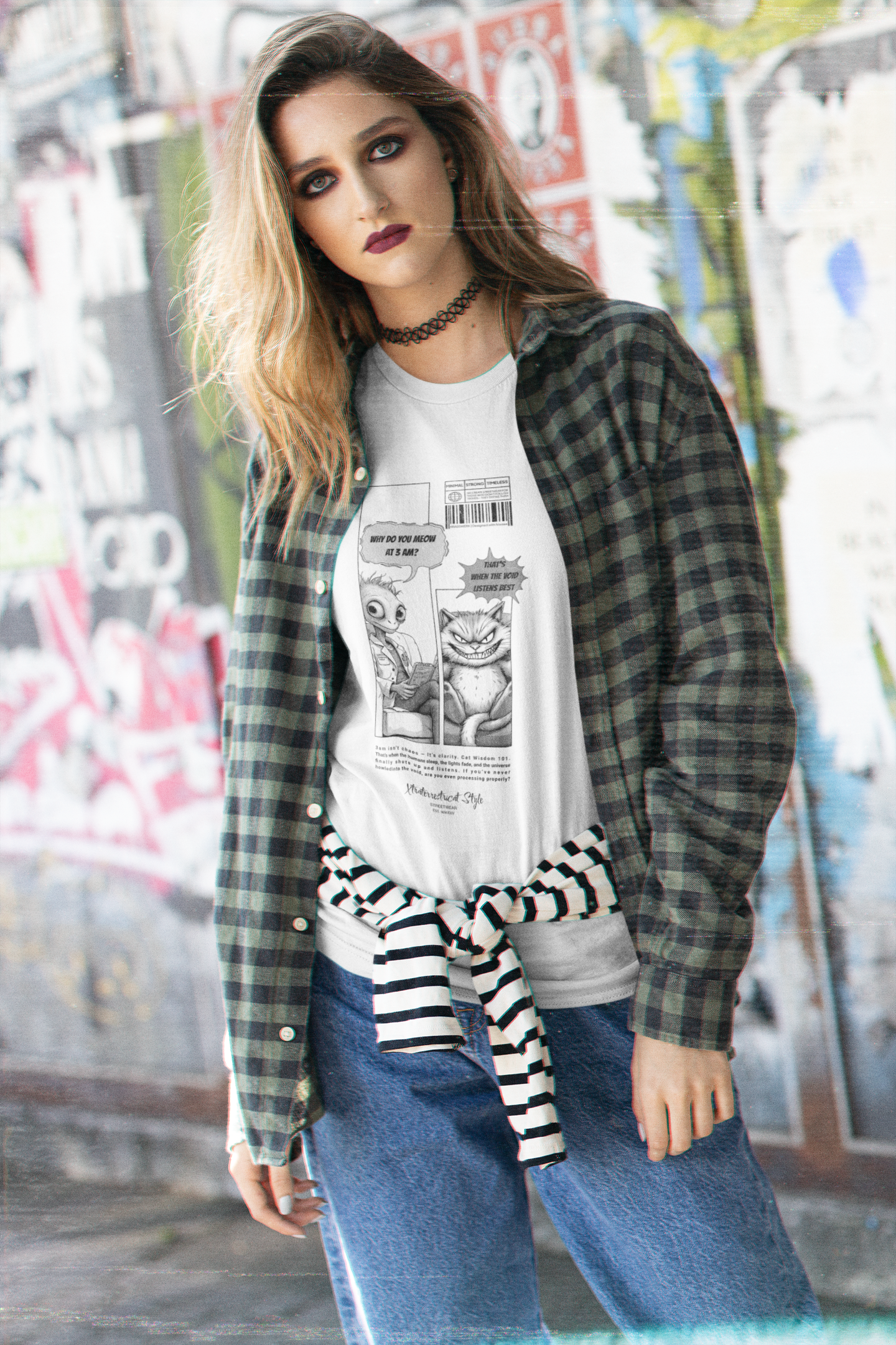 A young woman with bold makeup and loose wavy hair stands against a colorful graffiti wall, wearing a white t-shirt under a green plaid shirt. The t-shirt features a black-and-white comic strip with an alien asking, “Why do you meow at 3 A.M.?” and a smug cat replying, “That’s when the void listens best.” The shirt has a streetwear aesthetic, blending humor and urban style.