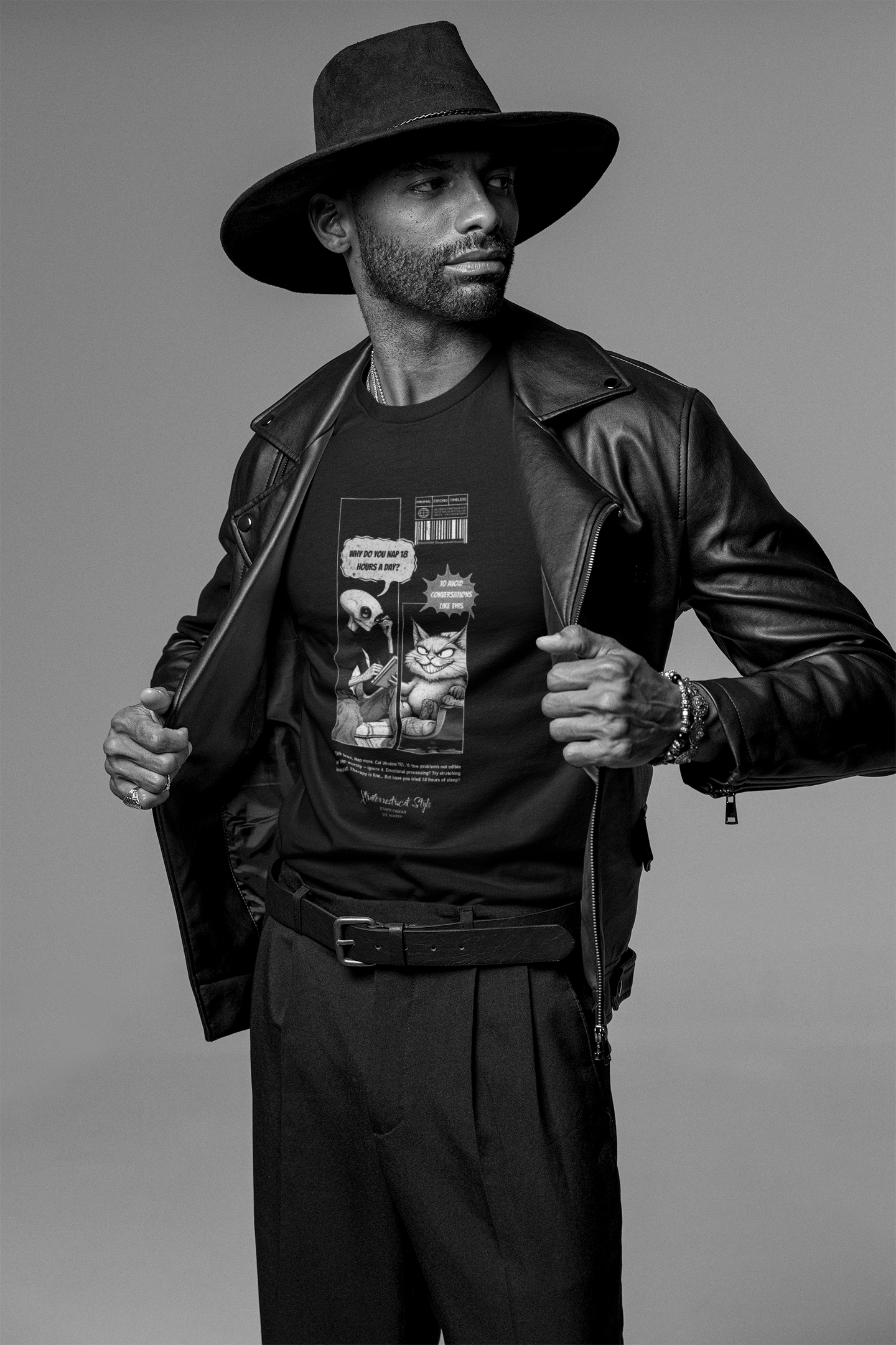 A confident male model in a wide-brim hat and leather jacket reveals a black t-shirt featuring a comic-style design of a cat and alien in therapy. The text reads: “Why do you nap 18 hours a day?” with the cat replying, “Some conversations take time.” Monochrome styling highlights the sarcasm and personality of the print.