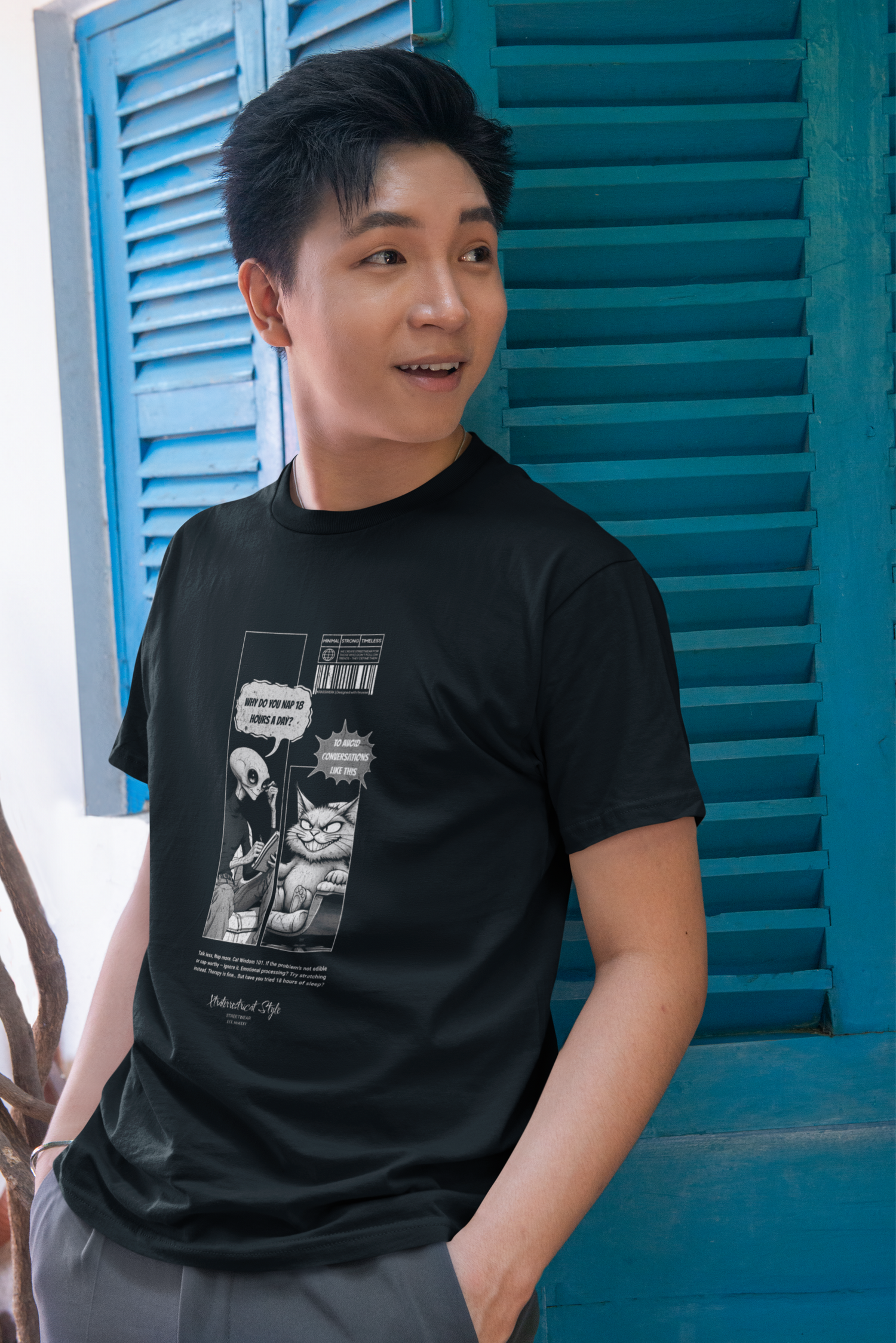 A relaxed man standing in front of bright blue shutters, smiling casually while wearing a black t-shirt featuring a sarcastic alien-cat comic. The comic shows an alien asking, “Why do you nap 18 hours a day?” and a grumpy cat responding, “Some conversations take time.” The monochrome print adds to the shirt’s humorous and quirky style.