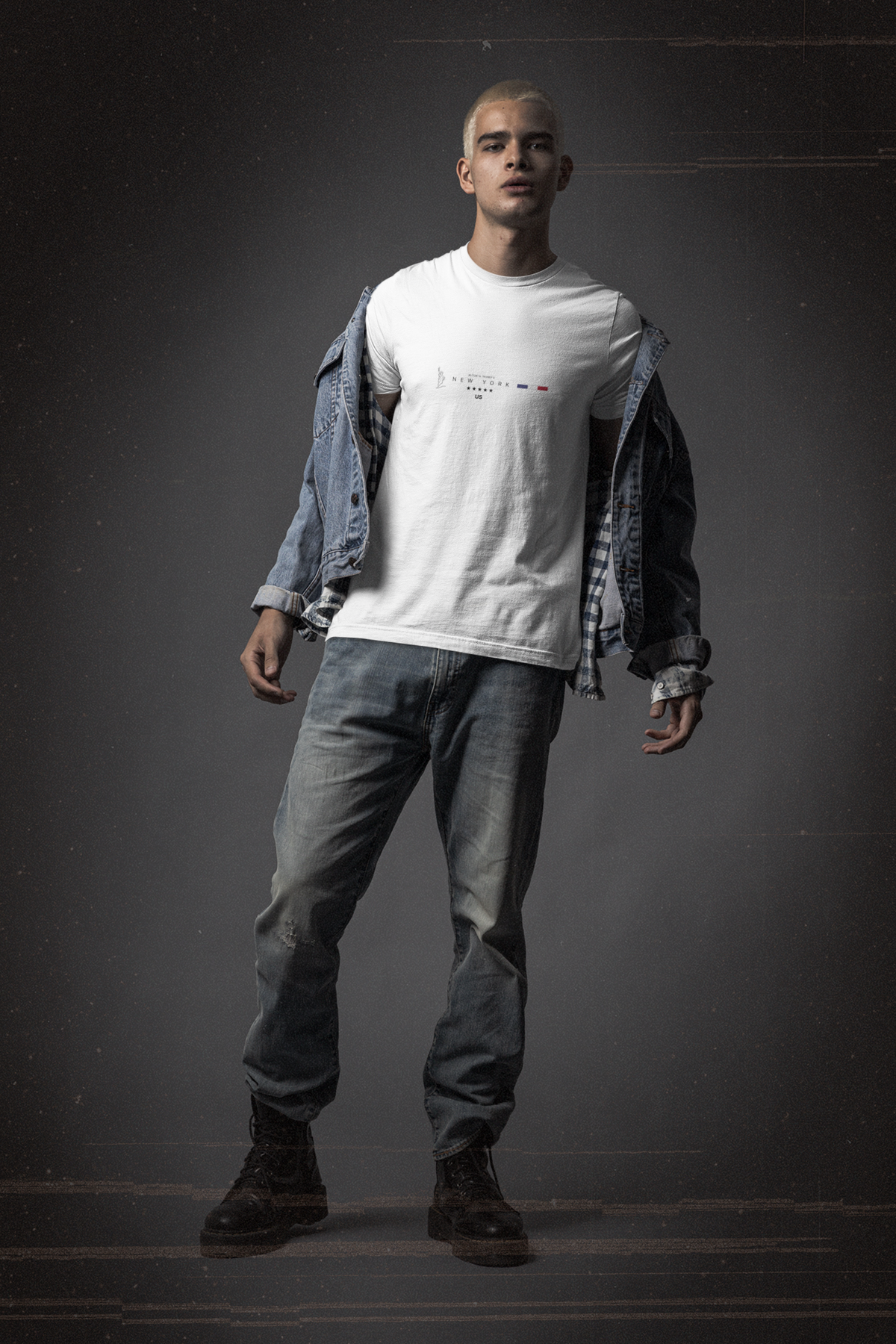 A young man stands confidently against a dark studio backdrop, wearing a white t-shirt with the NEW YORK design — featuring the Statue of Liberty icon, bold type, five stars, and a red-white-blue stripe. Styled with a half-open denim jacket and faded jeans, his pose and rugged boots convey urban grit and rebellious cool — a look that echoes the raw energy of New York City.