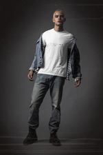 A young man stands confidently against a dark studio backdrop, wearing a white t-shirt with the NEW YORK design — featuring the Statue of Liberty icon, bold type, five stars, and a red-white-blue stripe. Styled with a half-open denim jacket and faded jeans, his pose and rugged boots convey urban grit and rebellious cool — a look that echoes the raw energy of New York City.