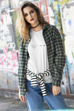A young woman poses confidently in front of a graffiti-covered urban wall. She wears a white t-shirt featuring the NEW YORK design — with the Statue of Liberty icon, bold text, five stars, and a stylized red-white-blue stripe. Layered with an open flannel shirt and striped sweater tied at the waist, the look blends streetwear and rebellion, echoing the spirit of NYC.