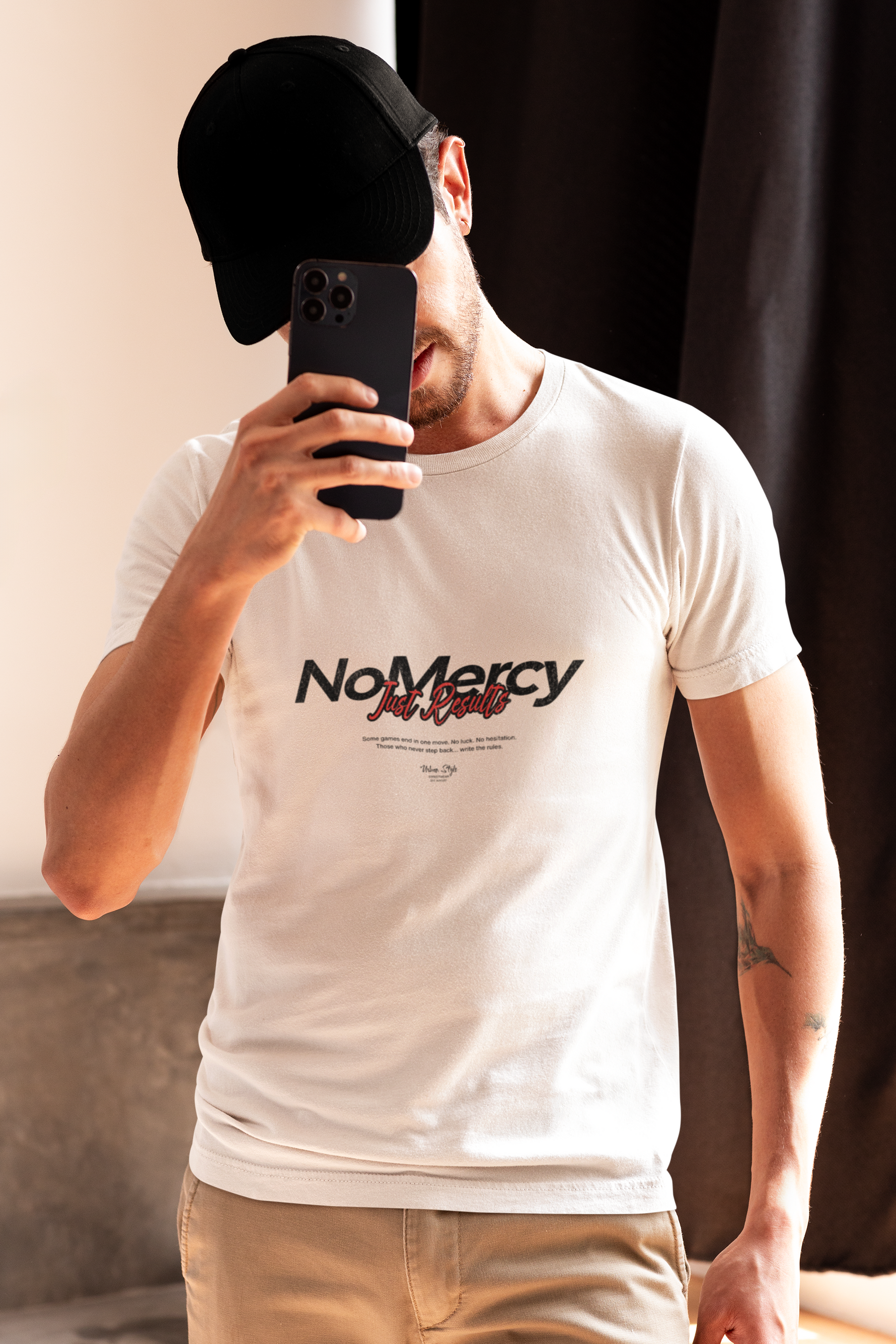 Man in a white NO MERCY – Just Results t-shirt taking a selfie indoors, wearing a black cap and beige pants. The bold black-and-red lettering stands out under warm lighting.
