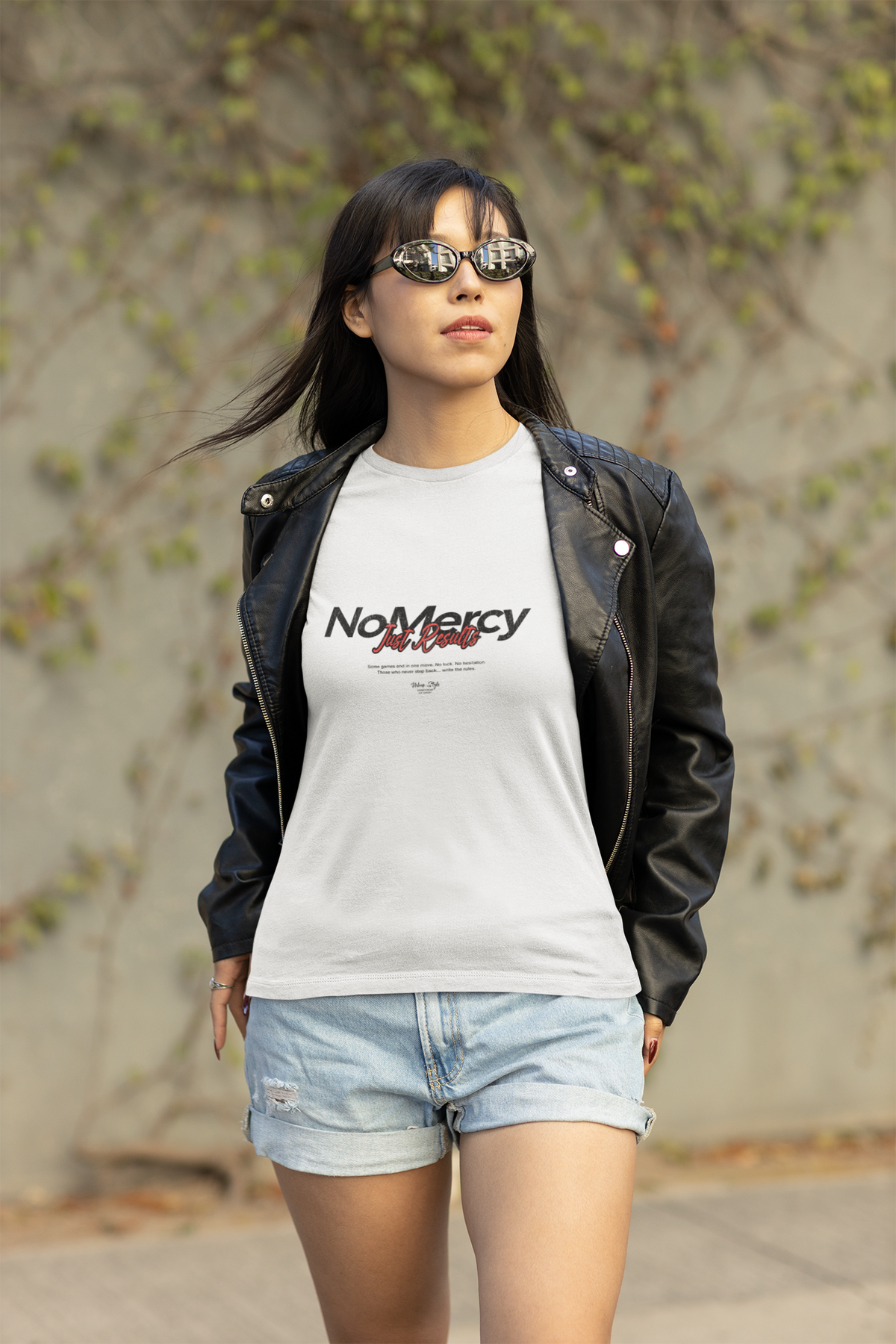 Confident woman walking outdoors in a leather jacket and sunglasses, wearing the NO MERCY – Just Results white t-shirt from Krasswerk. The bold black-and-red typography stands out as she moves with fearless intent.