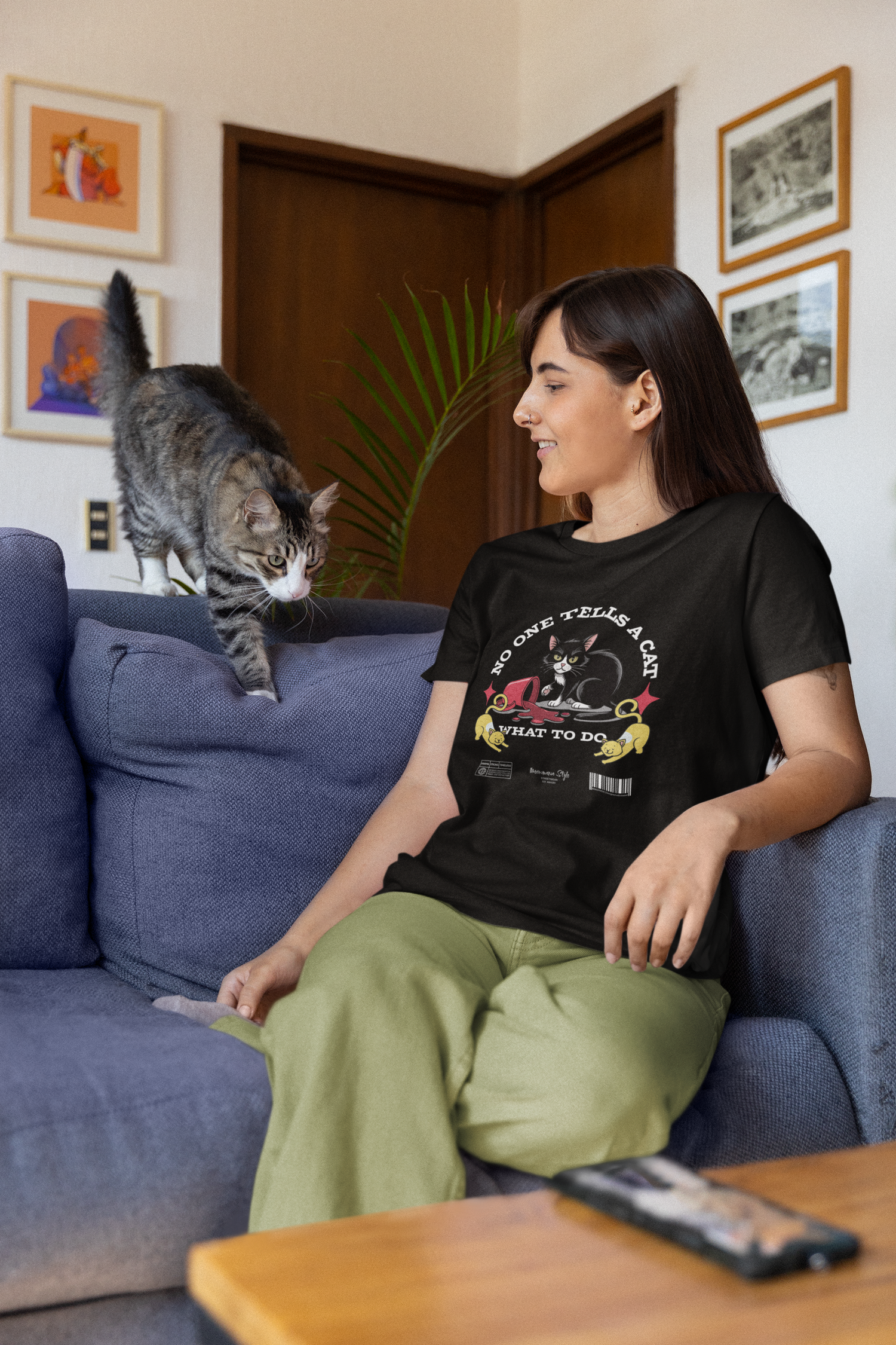 Woman sitting on a couch smiling at a curious cat, wearing a black “NO ONE TELLS A CAT WHAT TO DO” t-shirt featuring a rebellious cartoon cat with glasses and yellow hands pointing at it.