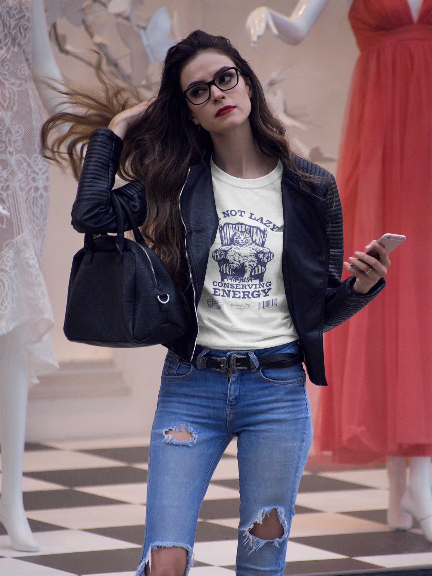 Woman with long brown hair flips it confidently while walking in a boutique mall. She wears ripped jeans, a leather jacket, and the white “NOT LAZY / I’m Just Conserving Energy” t-shirt featuring a lounging cat illustration. Stylish mannequins in the background complete the upscale setting.