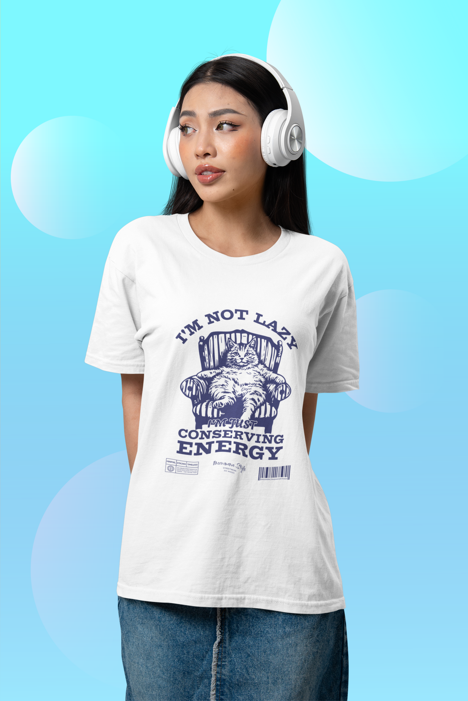 Young woman wearing large white headphones and a white “I’m Not Lazy / I’m Just Conserving Energy” t-shirt with a relaxed cat illustration. She poses against a playful blue background with circular shapes, looking off-camera with a thoughtful expression.