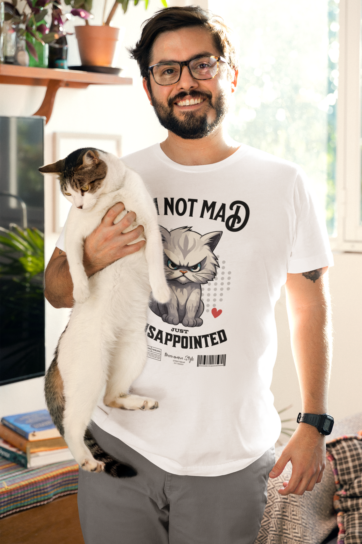 A bearded man wearing glasses stands smiling in a bright living room, holding a tall white cat against his chest. He’s wearing the NOT MAD white t-shirt, featuring an angry cartoon cat and the phrase “I’m not mad, just disappointed.” Sunlight pours through the window behind him, casting a cozy glow over the indoor plants and soft furnishings.