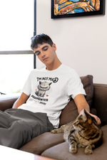 A young man sits casually on a brown sofa, gently petting a tabby cat resting beside him. He’s wearing a white t-shirt with a grumpy cartoon cat and the slogan “I’m not mad, just disappointed.” Soft daylight filters through a nearby window, and a colorful painting hangs on the wall above him. The calm, homey setting contrasts humorously with the shirt’s bold attitude.