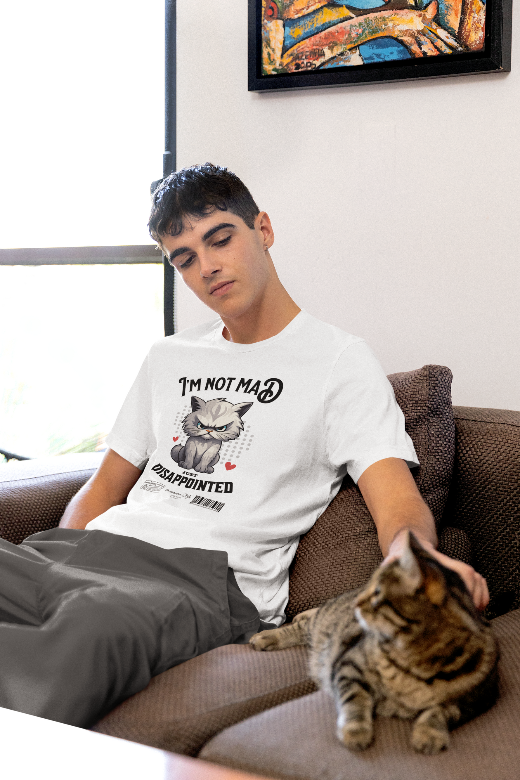 A young man sits casually on a brown sofa, gently petting a tabby cat resting beside him. He’s wearing a white t-shirt with a grumpy cartoon cat and the slogan “I’m not mad, just disappointed.” Soft daylight filters through a nearby window, and a colorful painting hangs on the wall above him. The calm, homey setting contrasts humorously with the shirt’s bold attitude.
