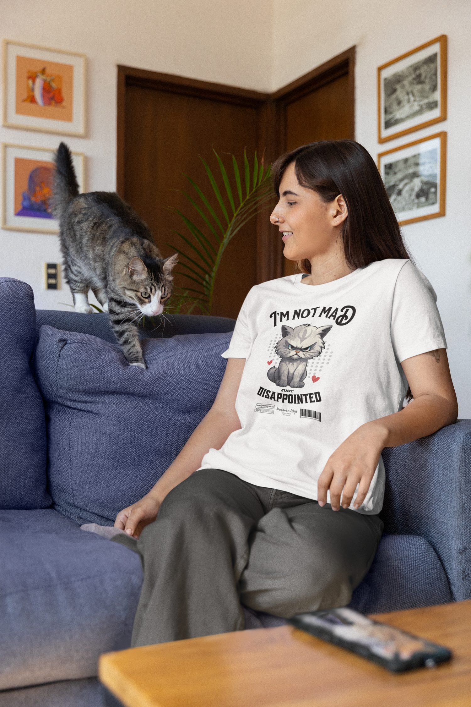 A young woman sits comfortably on a blue couch in a cozy living room, smiling as a curious tabby cat approaches her from the armrest. She wears the white NOT MAD t-shirt featuring an angry grey cartoon cat and the bold text “I’m not mad, just disappointed.” Framed art and a leafy plant fill the background, adding warmth and personality to the relaxed setting.