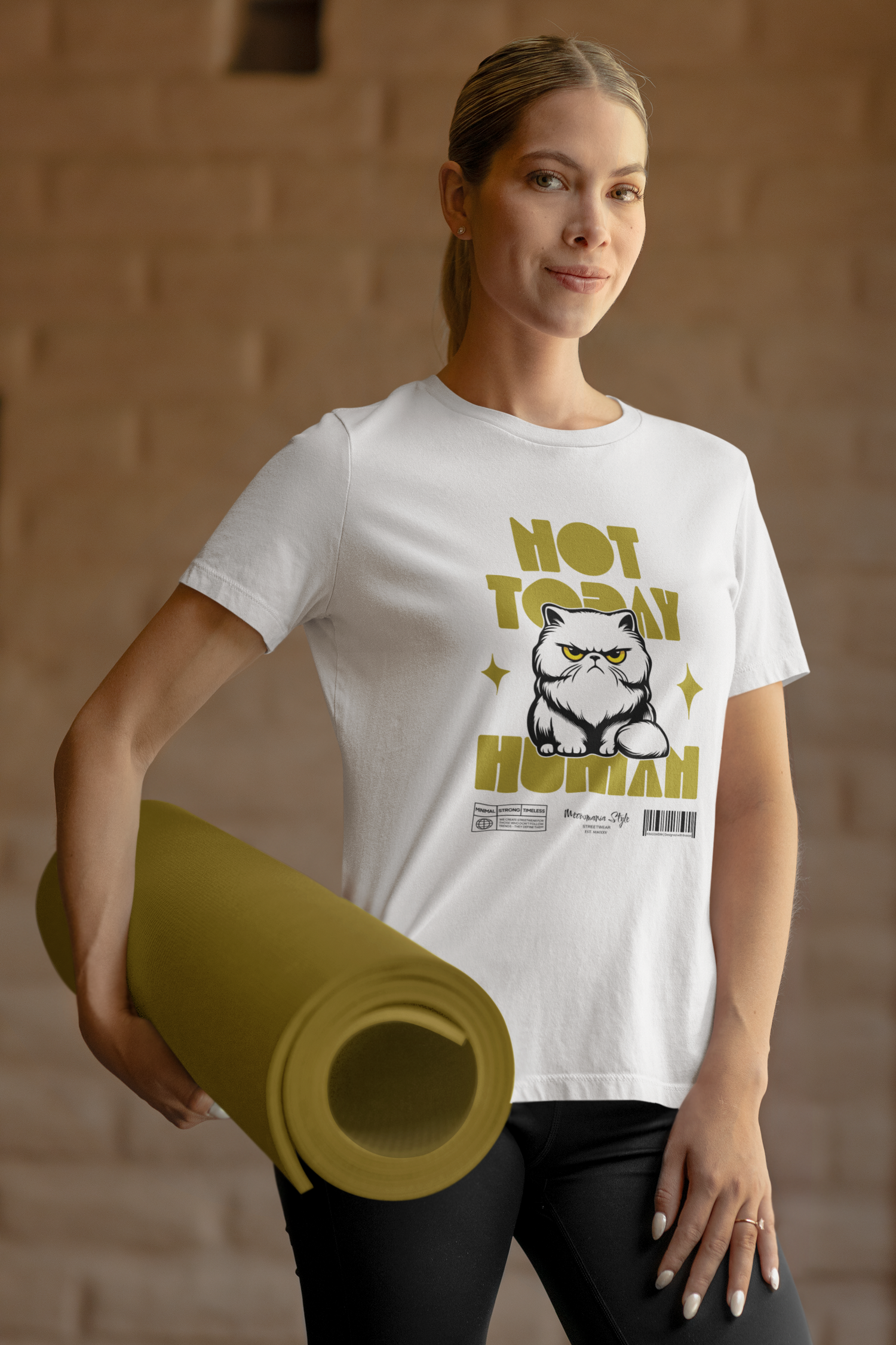 A woman standing confidently with a rolled olive-green yoga mat in one hand, wearing a white t-shirt printed with an angry Persian cat and the phrase ‘NOT TODAY HUMAN’ in bold yellow. She’s in a warmly lit interior with a beige stone wall, giving the scene a calm yet assertive tone that contrasts with the shirt’s defiant humor.