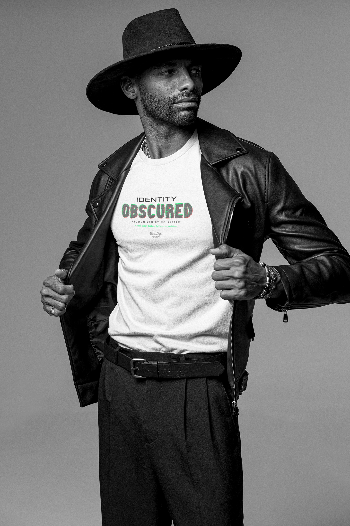 A confident man in a black leather jacket and wide-brimmed hat opens his coat to reveal a white t-shirt with the slogan “IDENTITY OBSCURED — RECOGNIZED BY NO SYSTEM.” Shot in black-and-white with sharp contrast. Studio background, no distractions.