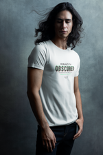 Man with long dark hair standing in front of a moody textured wall, wearing a white “IDENTITY OBSCURED” t-shirt with glitch-style green and red text. His eyes are closed, giving the scene a calm, introspective feel.