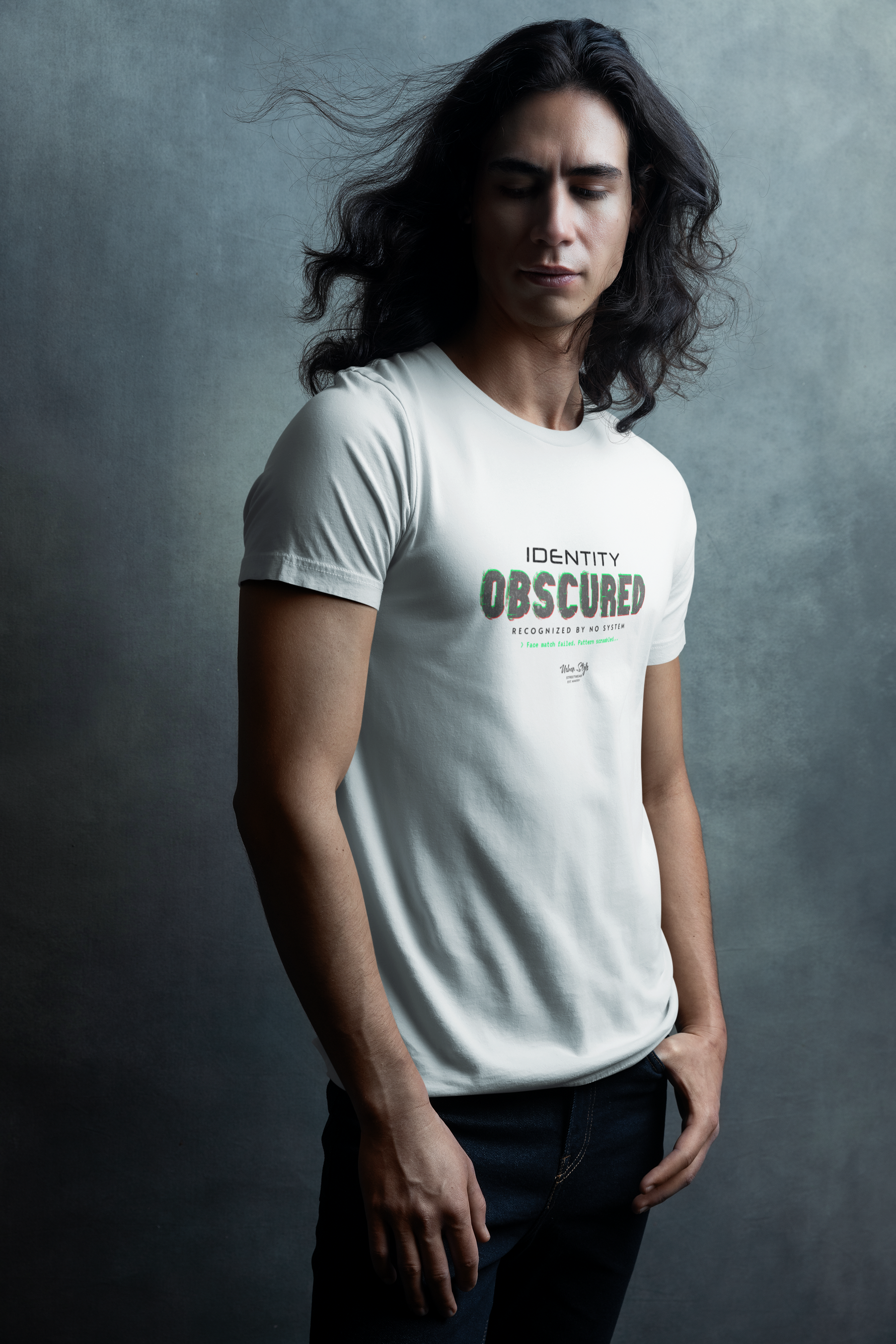 Man with long dark hair standing in front of a moody textured wall, wearing a white “IDENTITY OBSCURED” t-shirt with glitch-style green and red text. His eyes are closed, giving the scene a calm, introspective feel.