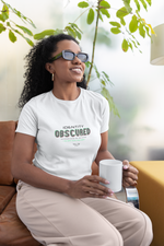 Woman sitting on a sofa in a relaxed setting, smiling and holding a white mug. She wears a white “IDENTITY OBSCURED” t-shirt with glitch-style green and red text. Indoor background with soft natural light and green plants.