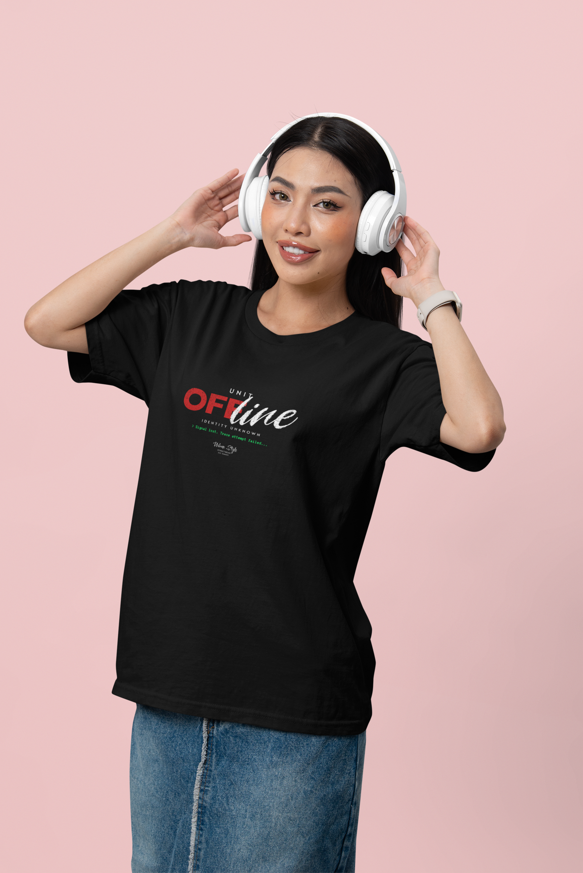 A woman in a black “UNIT OFFLINE” T-shirt poses against a soft pink background, adjusting her white over-ear headphones with a relaxed smile. The shirt design features bold, glitch-style typography in red, white, and green.