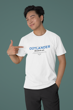 Male model smiling and pointing at the white OUTLANDER t-shirt — bold blue “Built outside the system” slogan stands out on a minimalist teal background.