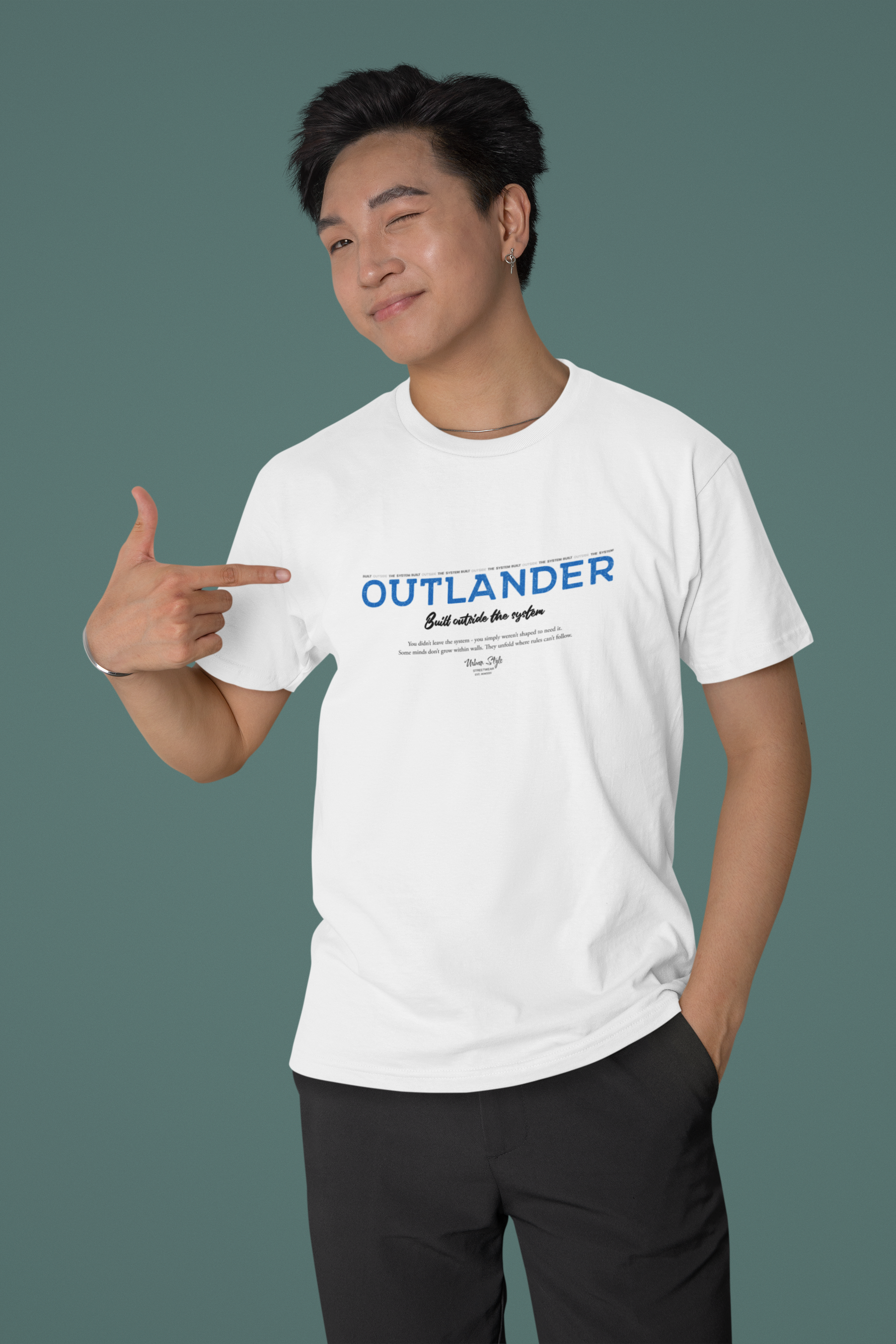 Male model smiling and pointing at the white OUTLANDER t-shirt — bold blue “Built outside the system” slogan stands out on a minimalist teal background.