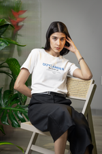 Female model seated on a wooden chair in a minimal interior, wearing the white OUTLANDER t-shirt — blue “Built outside the system” print clearly visible above black skirt.