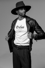 A bold man in a wide-brim hat and leather jacket reveals his "Outlaw – Make Your Own Path" T-shirt, standing with quiet defiance in a minimalist studio setting.