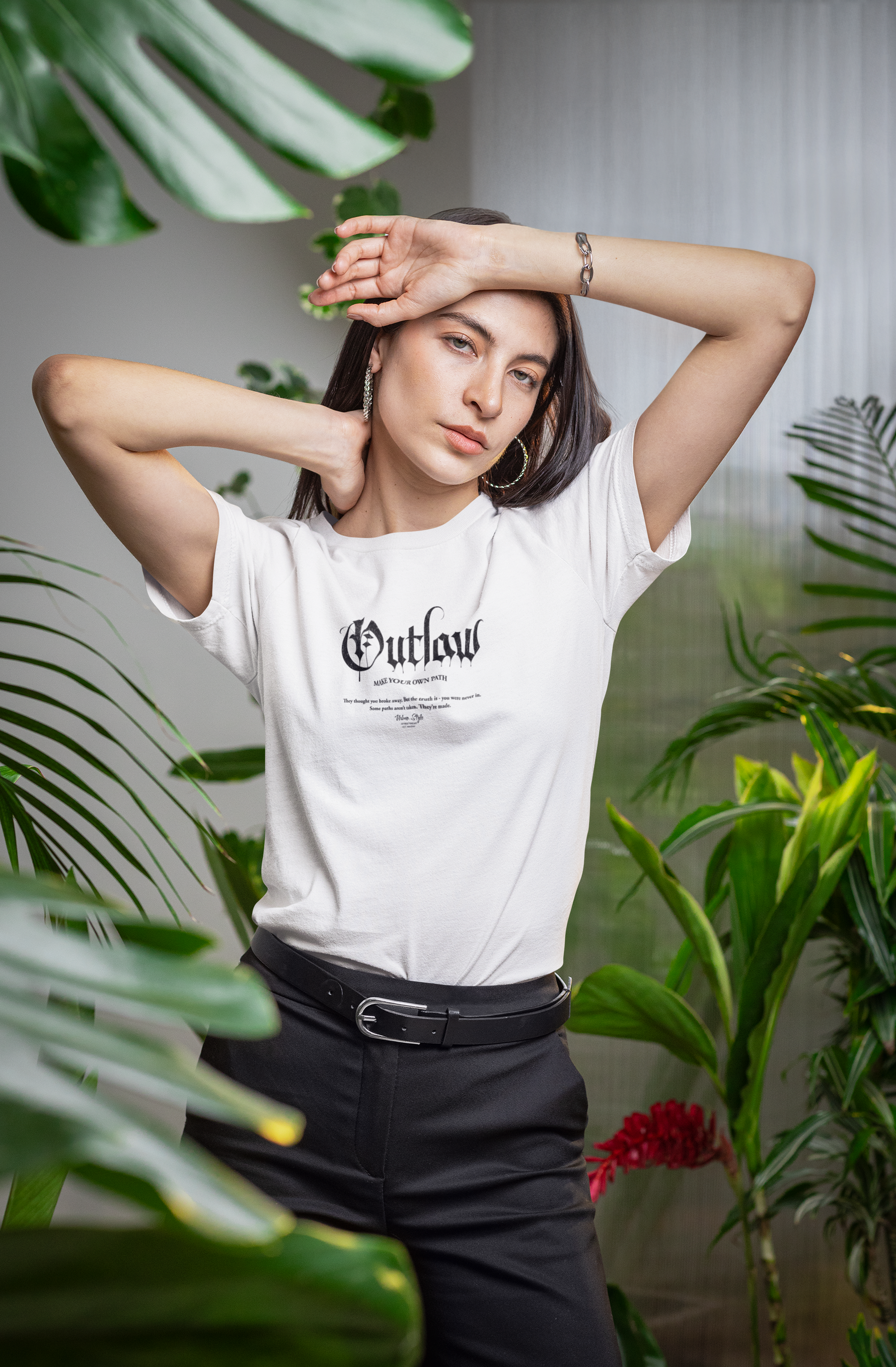 A confident woman stands among lush indoor plants, striking a bold pose in her "Outlaw – Make Your Own Path" white T-shirt, blending softness with rebellious energy.