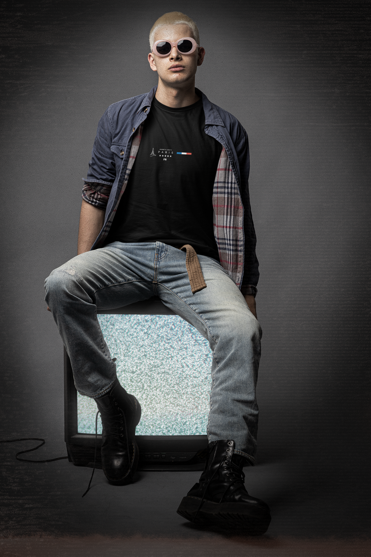 A bold male model in sunglasses and boots sits on a vintage static TV wearing the black PARIS t-shirt. The design features minimalist text, five stars, and a French flag bar, evoking Parisian edge with rebel energy. The setting is moody and stylish, matching the urban aesthetic.