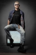 A bold male model in sunglasses and boots sits on a vintage static TV wearing the black PARIS t-shirt. The design features minimalist text, five stars, and a French flag bar, evoking Parisian edge with rebel energy. The setting is moody and stylish, matching the urban aesthetic.