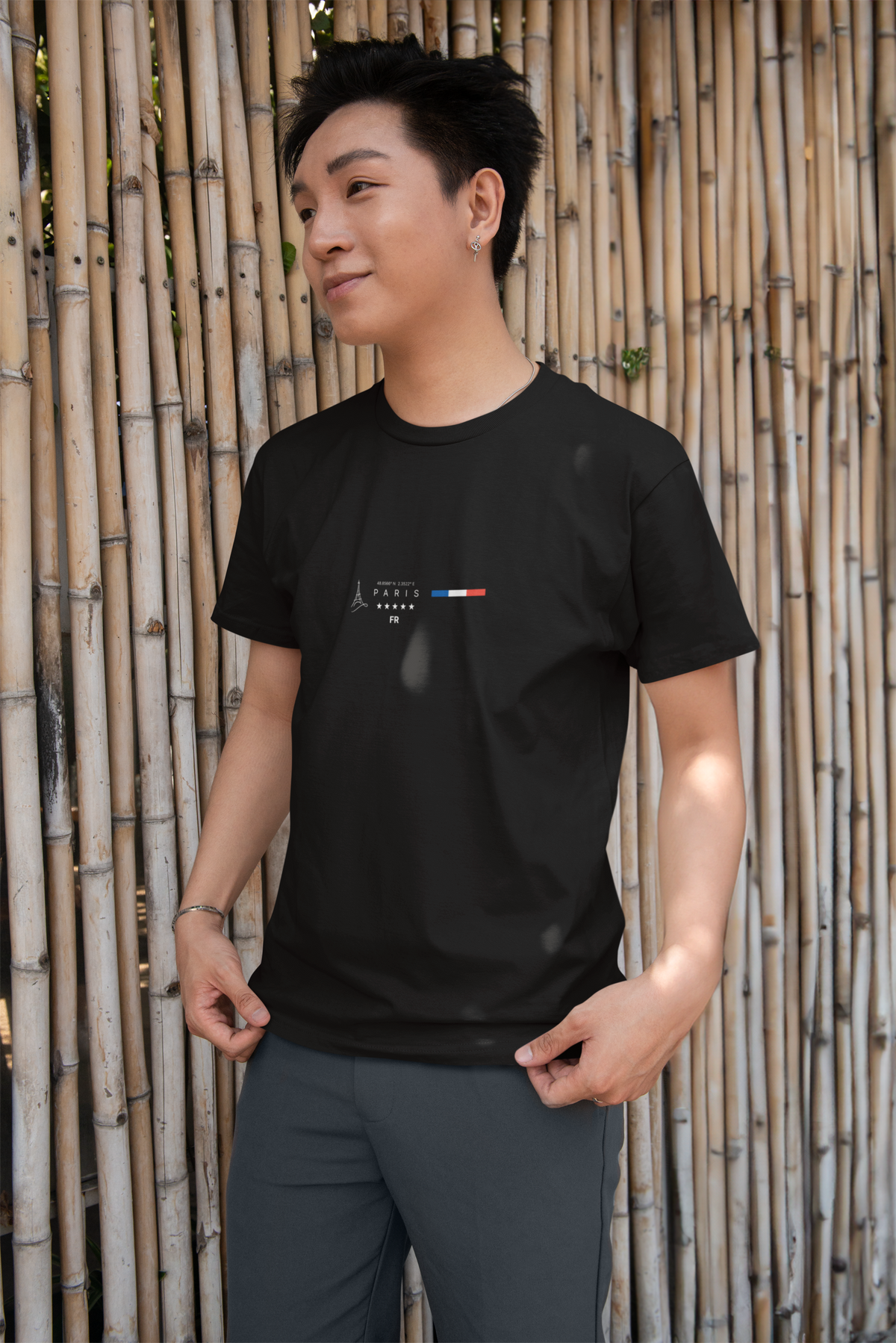 A man stands casually in front of a bamboo wall, wearing the black PARIS t-shirt. The design features the Eiffel Tower icon, bold Paris lettering, and a horizontal French flag motif. Natural light casts soft shadows over his shirt, blending urban style with a calm, outdoor vibe.
