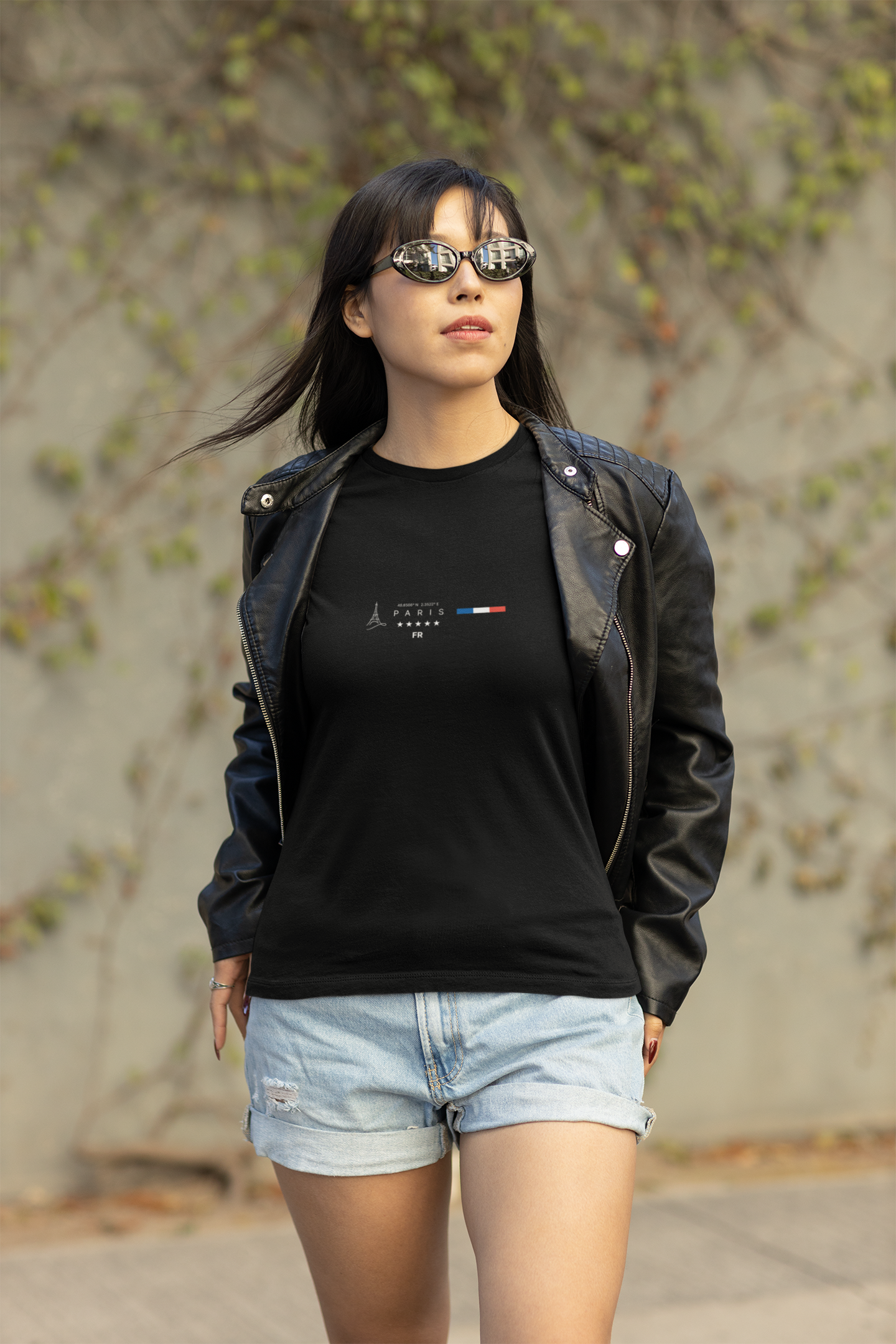 A confident woman walks outdoors in sunglasses and a leather jacket, wearing the black PARIS t-shirt. The minimalist design includes the Eiffel Tower, clean lettering, and French flag colors. Natural sunlight and urban vines in the background add contrast to her sharp streetwear vibe.