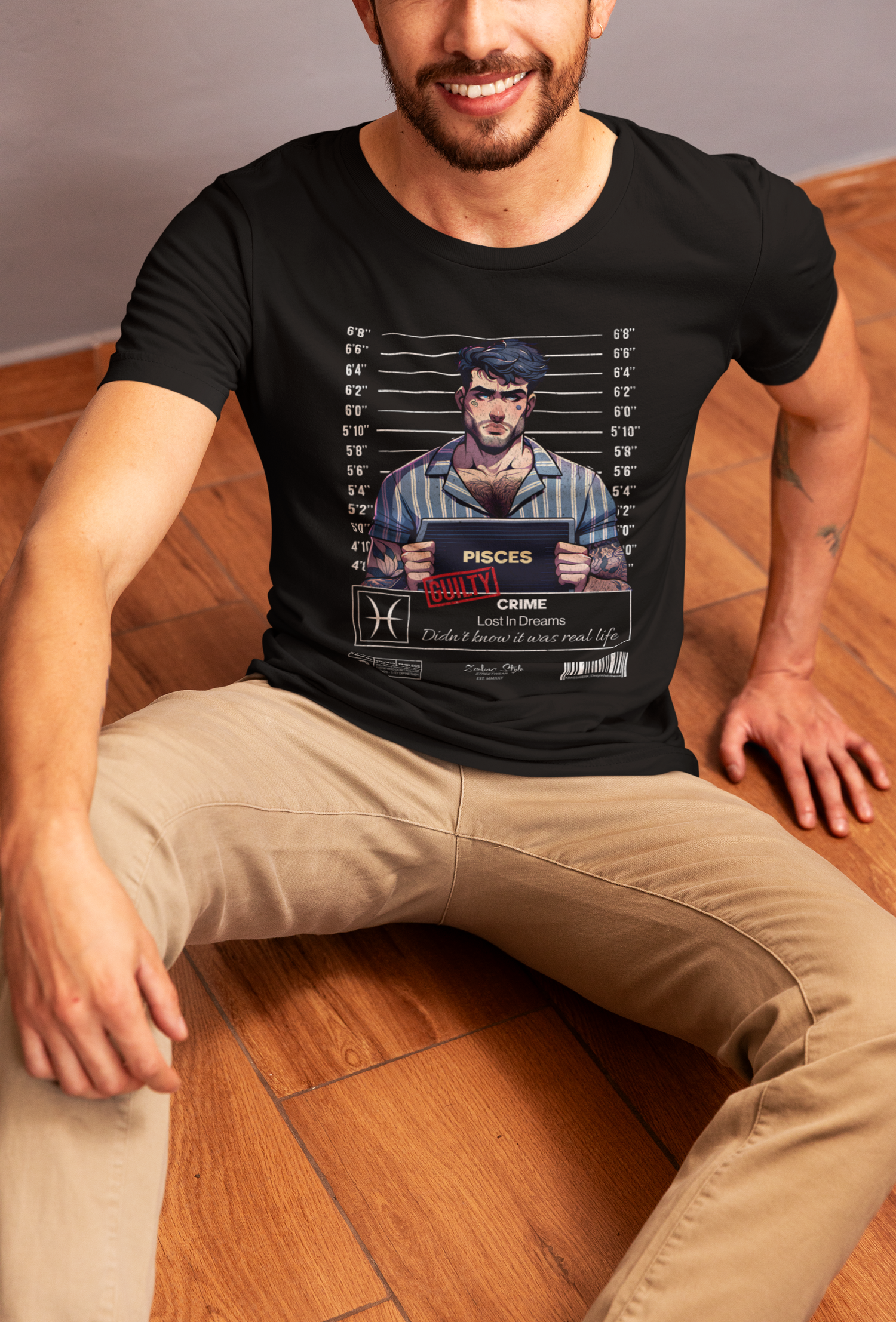 A man sits on a wooden floor, smiling slightly, wearing a black t-shirt with a colorful “PISCES – Lost In Dreams” mugshot design. The illustration features a dreamy-eyed man in striped pajamas holding a nameplate, with white height lines in the background.