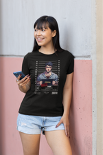 A woman stands casually against a pastel pink and concrete wall, smiling while holding a phone. She wears a black t-shirt featuring the “PISCES – Lost In Dreams” mugshot artwork, showing a dreamy-looking man in striped pajamas holding a nameplate with height lines behind him.